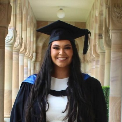 A woman with long black hair is smiling at the camera, she is wearing a graduation cap and gown over a white dress and is standing in the sandstone cloisters of UQ's Great Court.